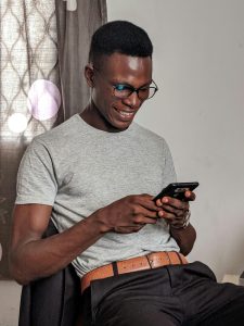 A happy young man uses his smartphone while sitting indoors, expressing joy.