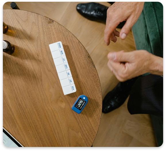 Medication Management at Regions Praxis Care – Safe, Personalized Support Close-up of a weekly pill organizer and pulse oximeter on a wooden table next to a seated person’s hands.